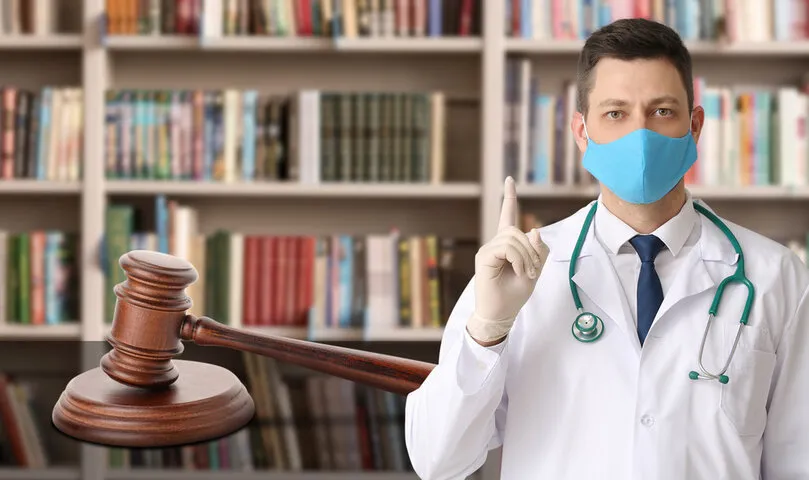 Male doctor in medical mask and with judge's gavel in library